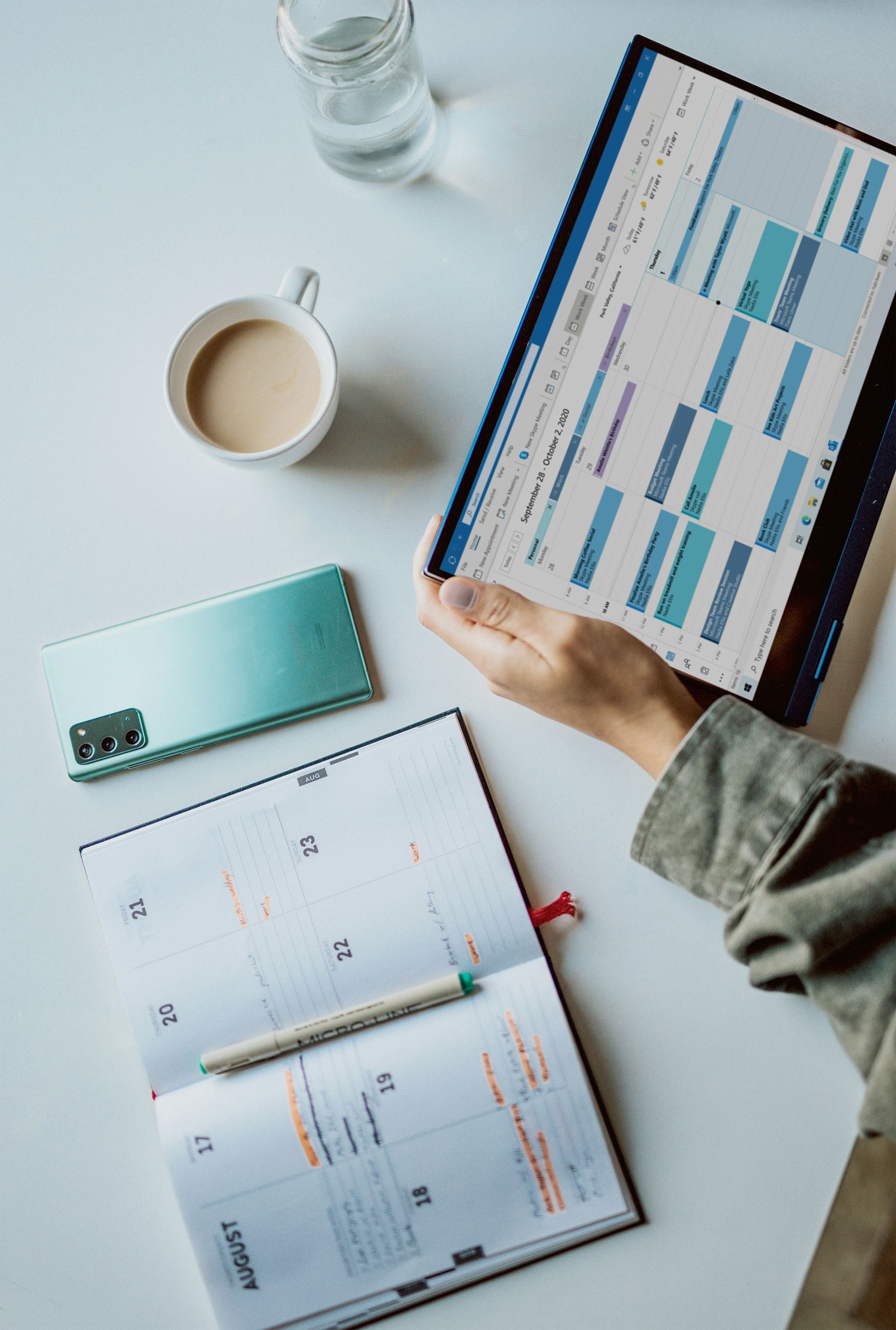 a person stood at a table holding a mobile tablet with an electronic calendar on the screen, a diary, a mobile phone and a cup of coffee is on the table. 